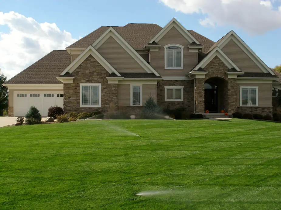 Stone and stucco home with sprinklers running across a wide front lawn