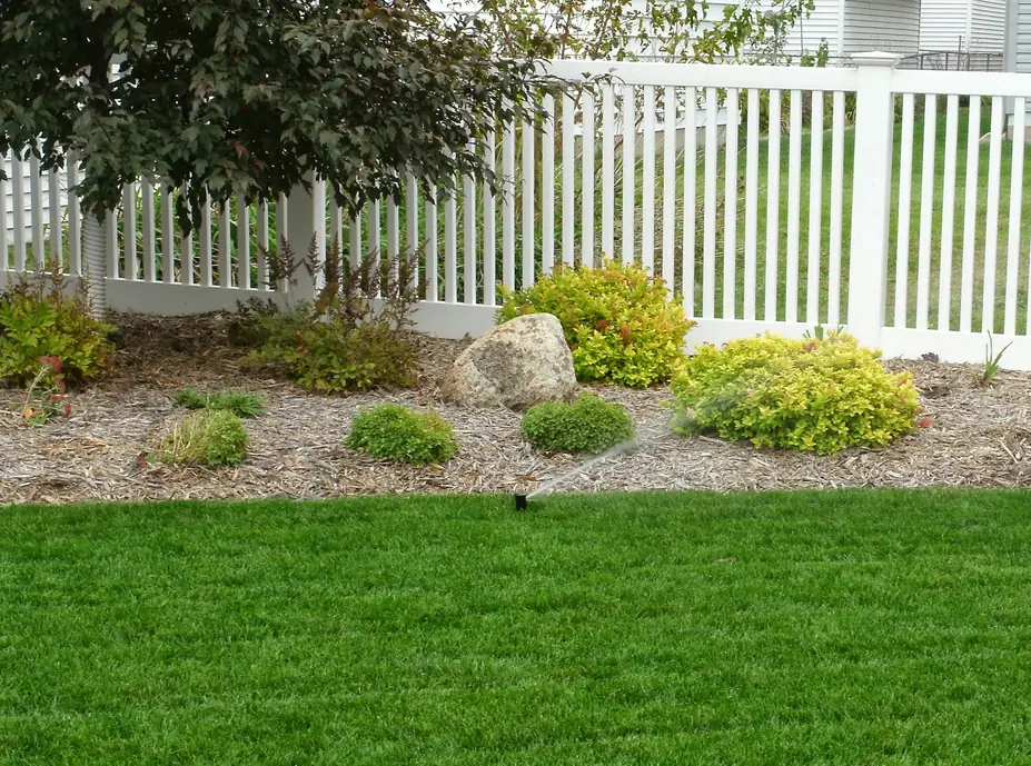 Planted bed along a white vinyl fence with a sprinkler head at the lawn edge